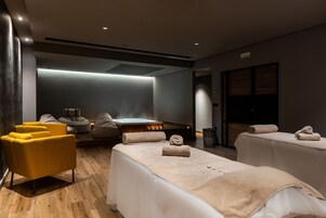 A modern spa room with wooden flooring, a yellow armchair, two massage tables with towels, a sauna, and a wooden bench.