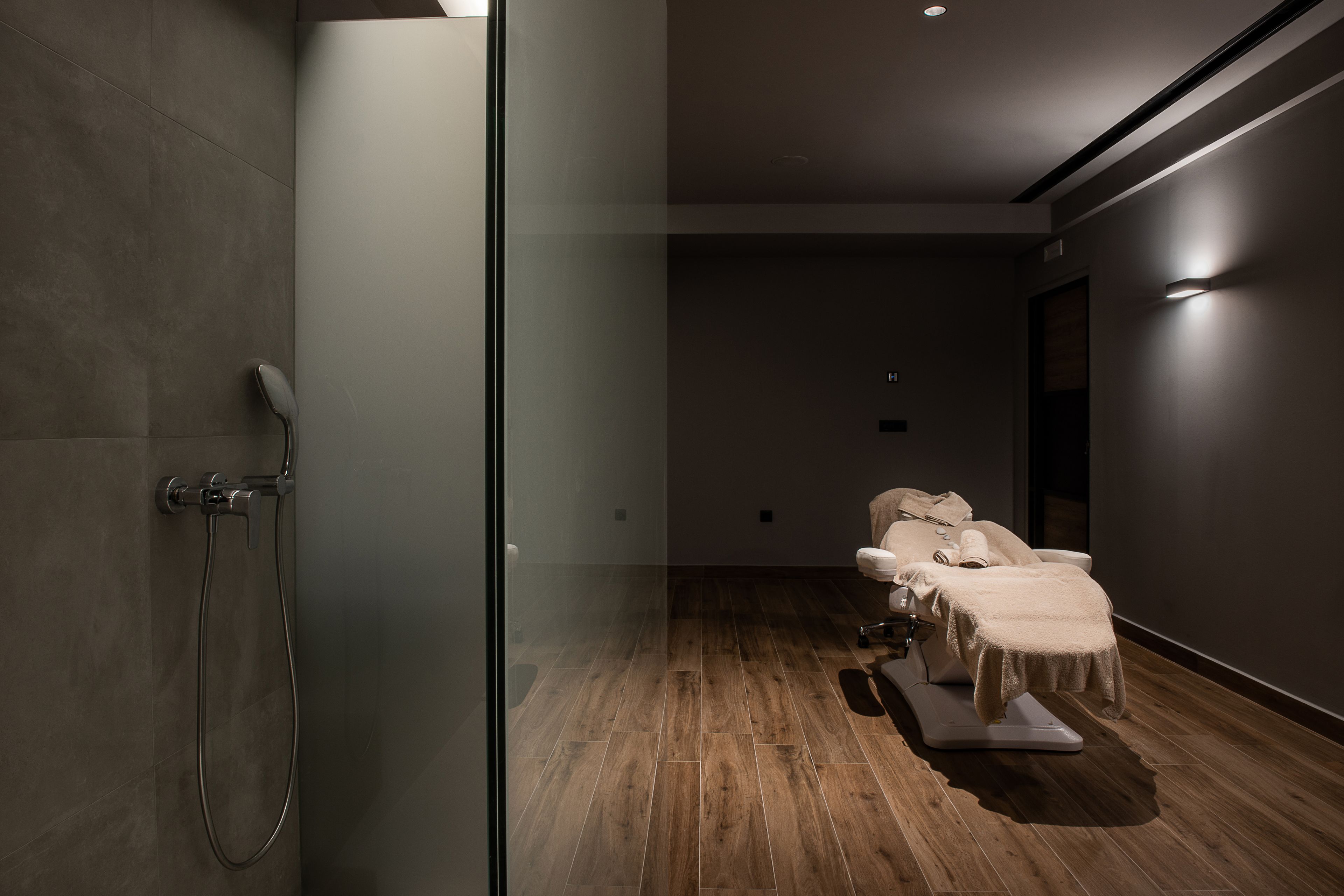 A modern spa room with a glass shower enclosure, wooden flooring, and a massage table with towels.