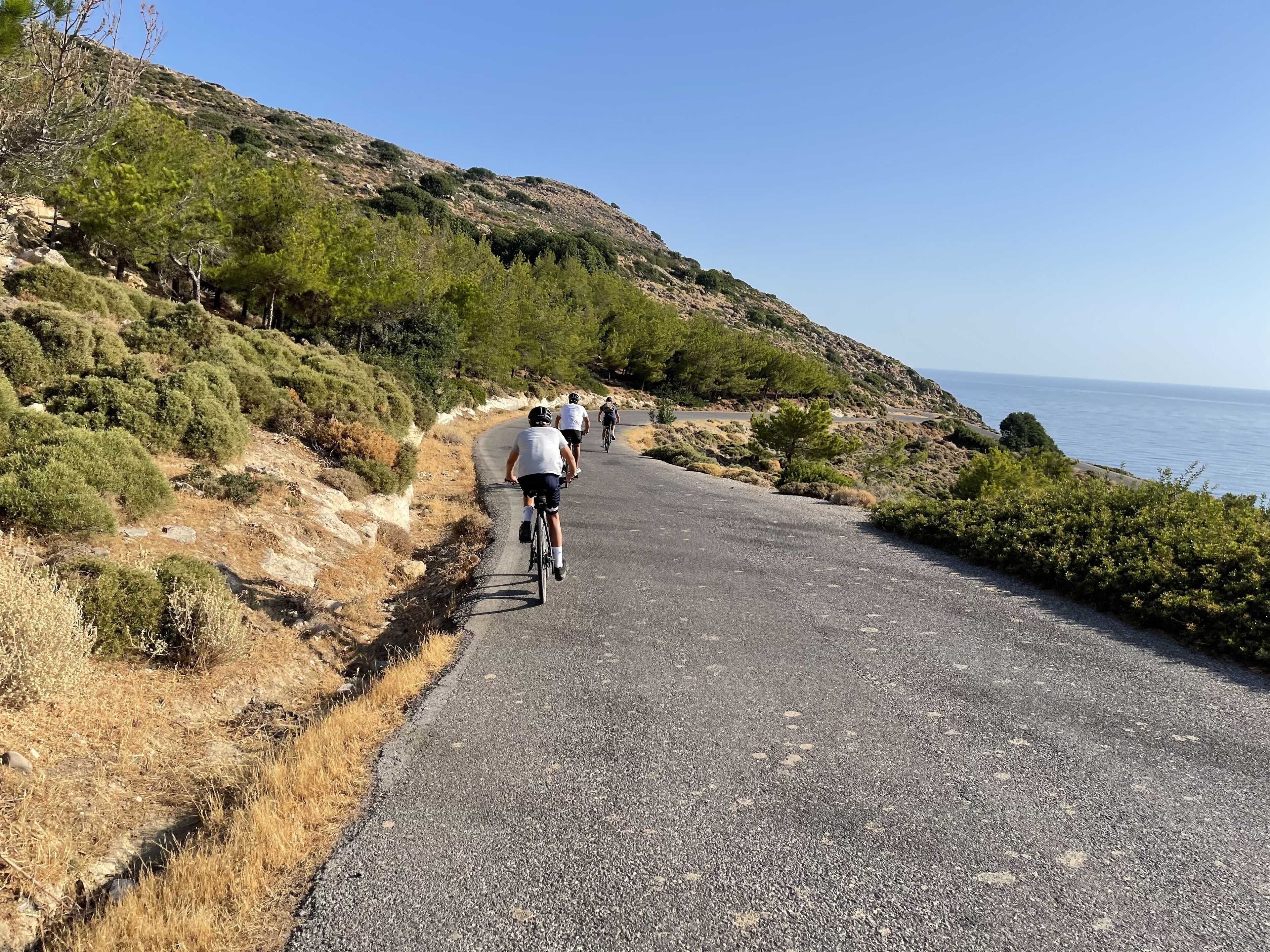 Una carretera pavimentada con ciclistas, rodeada de hierba seca y vegetación escasa, que conduce hacia una masa de agua.