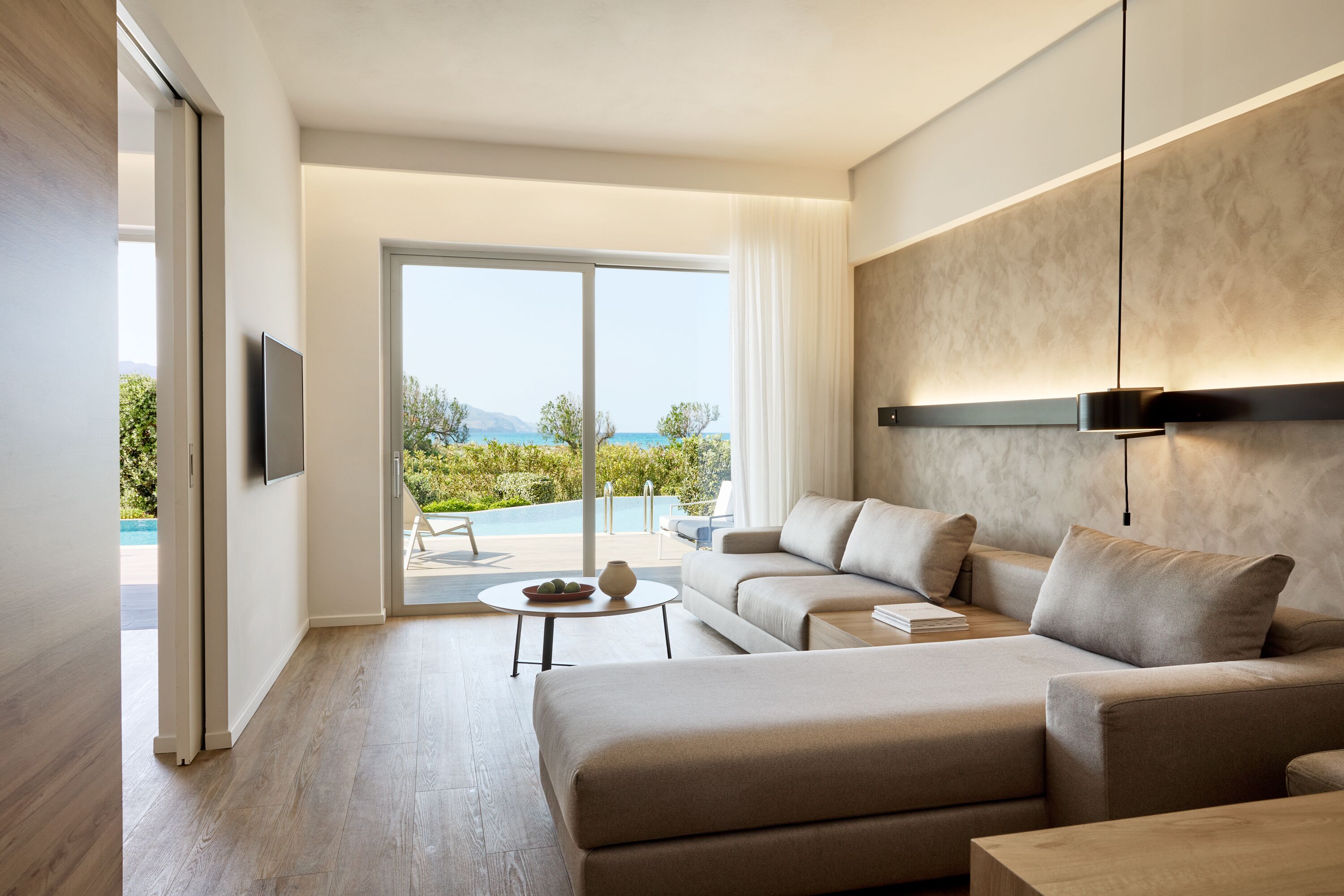 Una sala de estar moderna con un sofá, una mesa de centro y un televisor. Se ve una ventana grande con vista a una piscina y follaje.