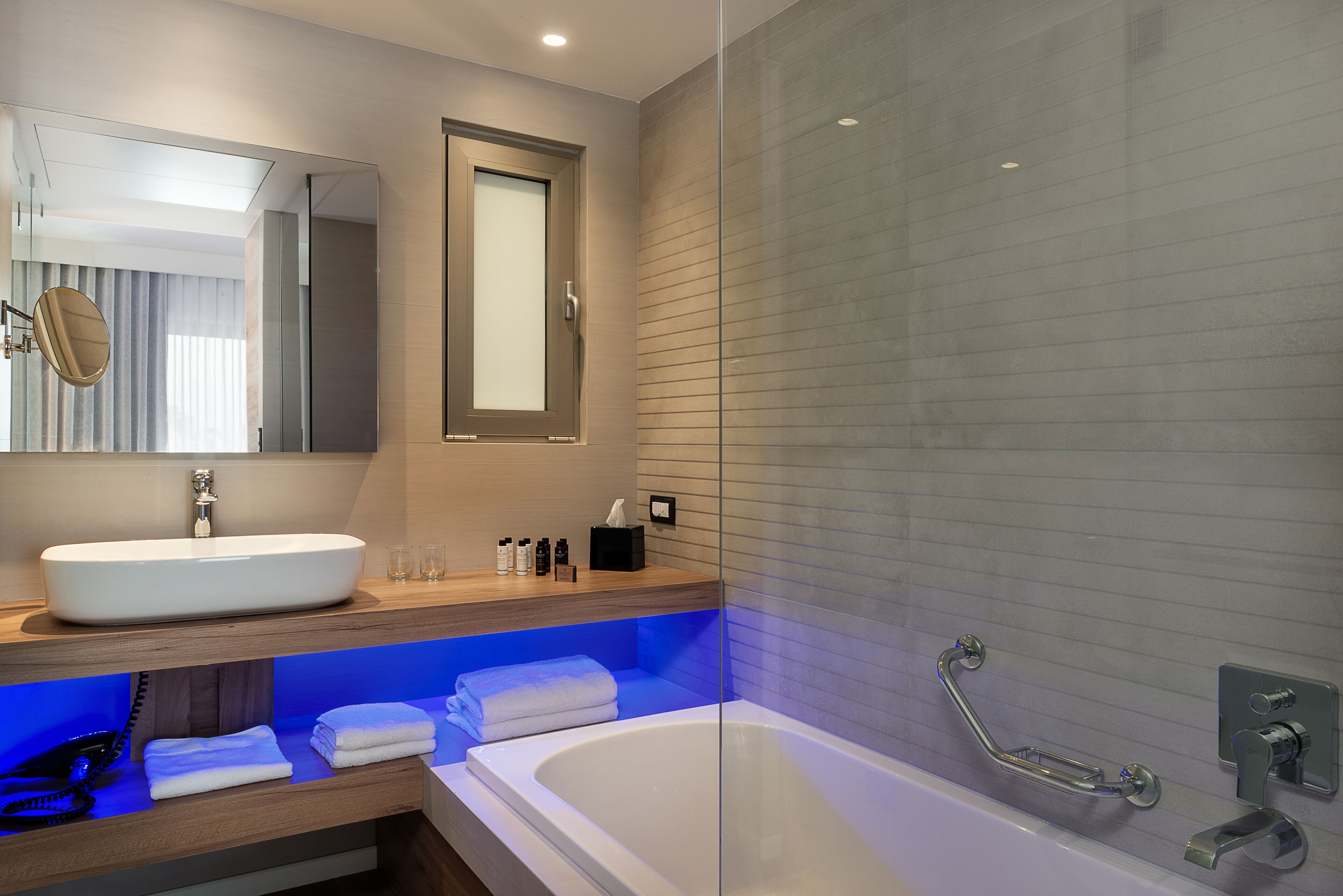 Un baño moderno con una bañera blanca grande, una ducha con mampara de vidrio y un tocador de madera con espejo y lavamanos.