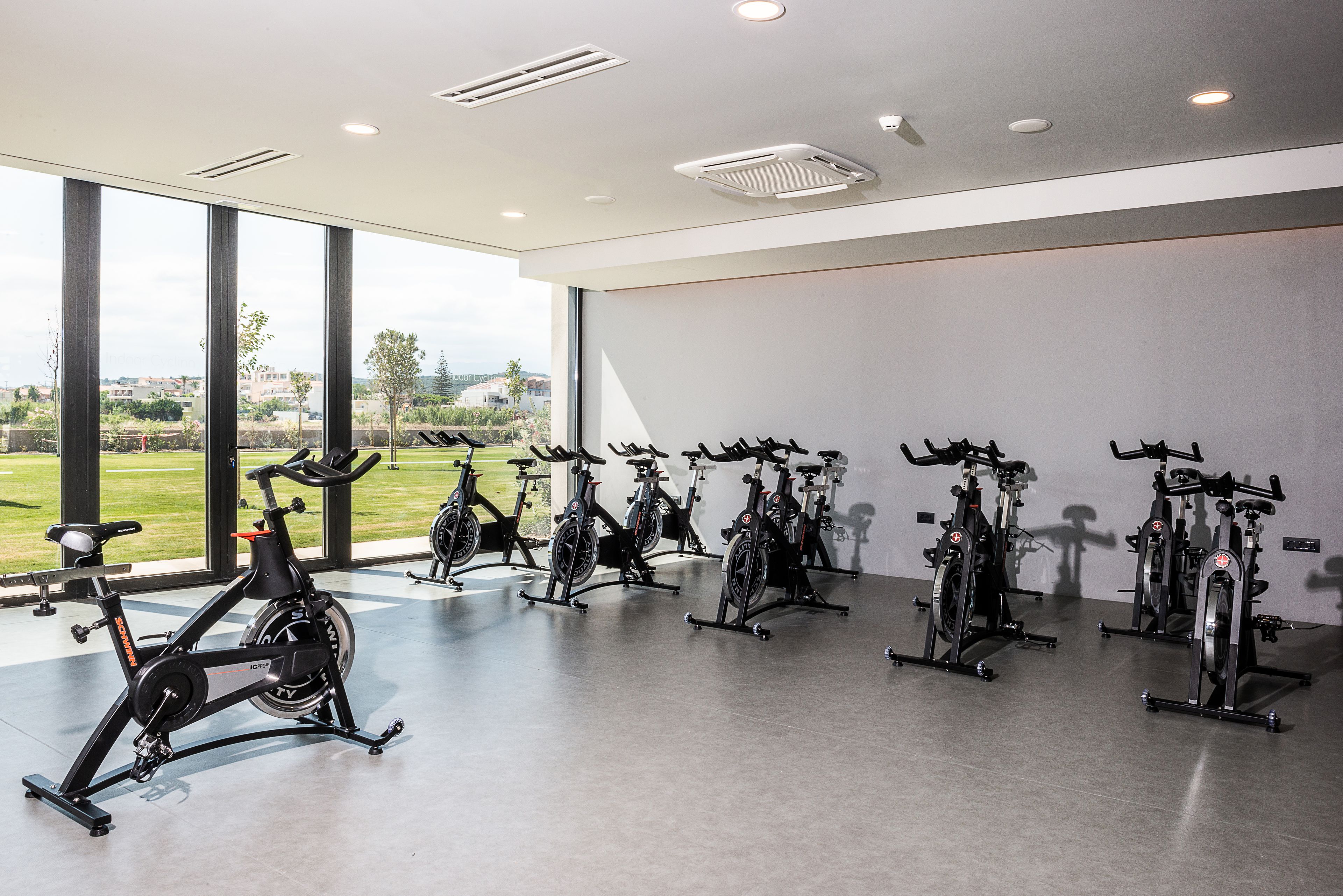 A gym with stationary bikes arranged in a row, large windows with a view of greenery and buildings, and a modern ceiling with recessed lighting.