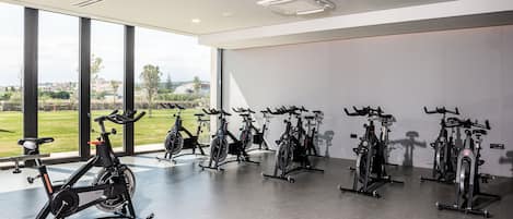 Ein Fitnessraum mit in Reihe angeordneten Ergometer-Rädern, großen Fenstern mit Blick auf Grünflächen und Gebäude sowie einer modernen Decke mit Einbauten für indirekte Beleuchtung.