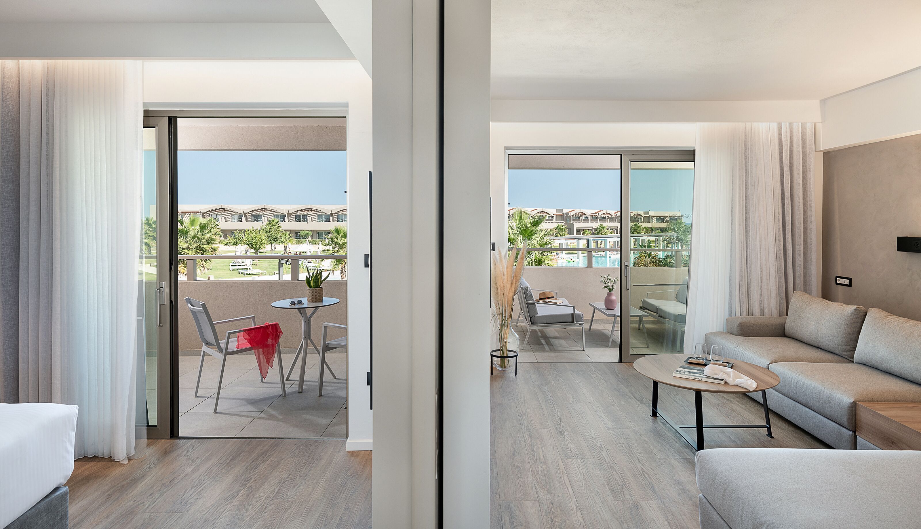 A modern hotel room with a balcony, a sofa, a coffee table, and a dining area with a table and chairs.