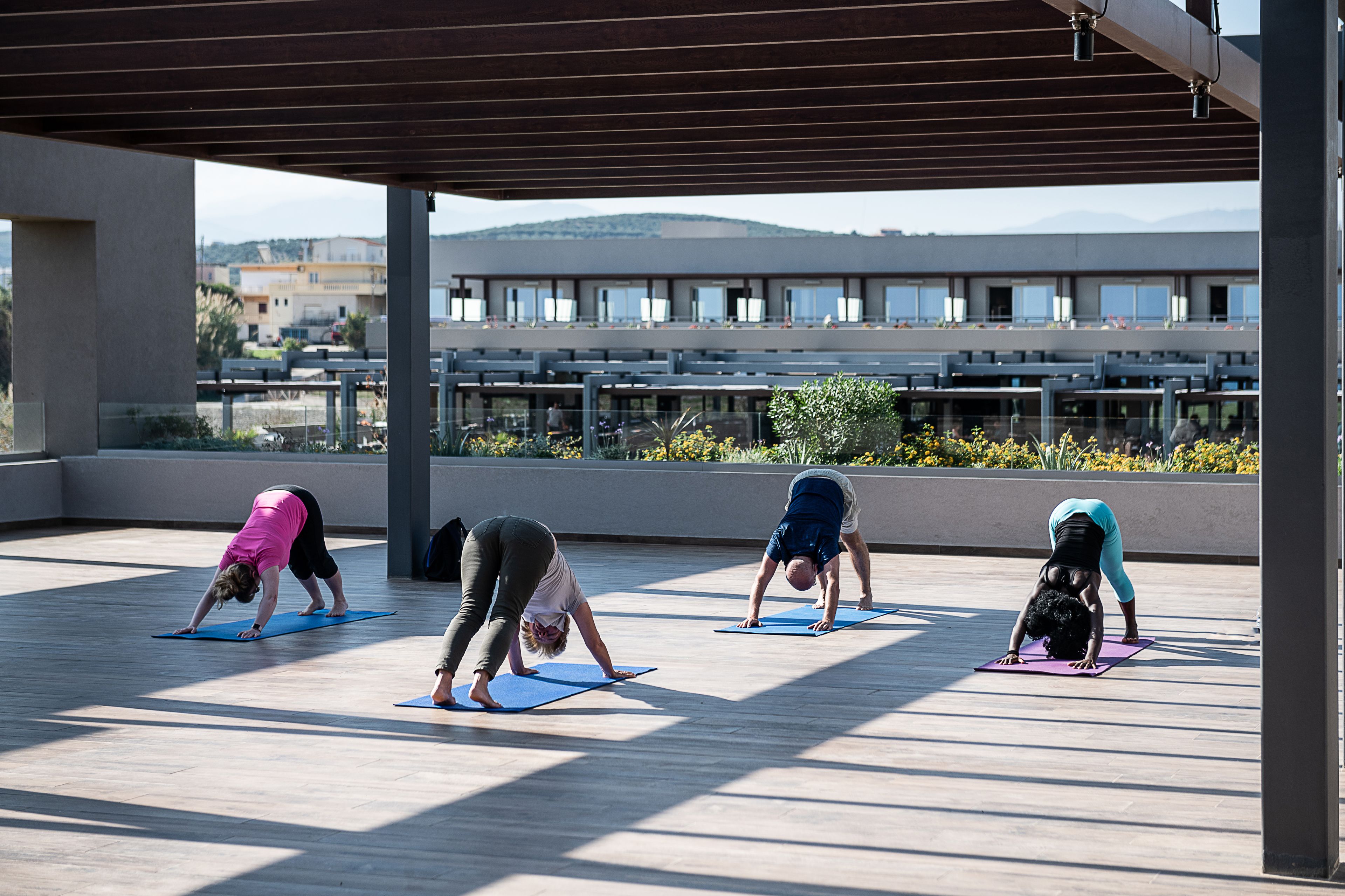 Four people doing yoga on blue mats under a covered area with a view of buildings and mountains in the background.