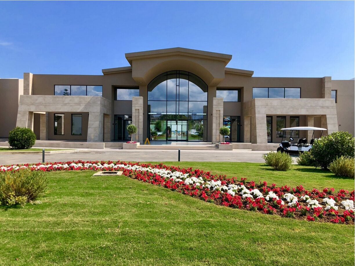 A modern building with a large entrance, a parking area, and a well-maintained garden with red and white flowers.