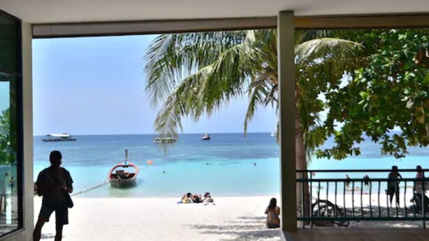 A beach view from inside a building with a glass wall, palm trees, and a boat anchored near the shore.