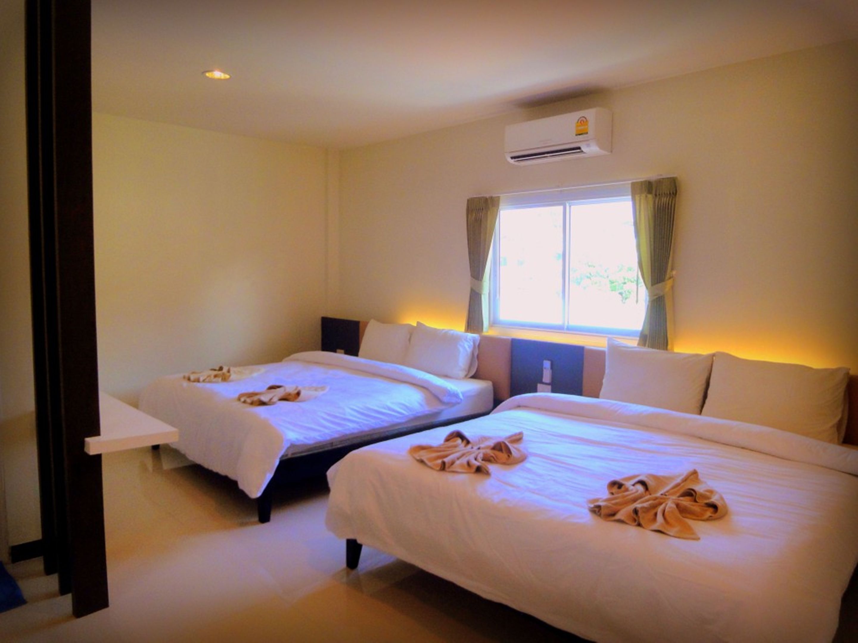 A hotel room with two beds, each with neatly folded towels. There is a window with curtains, an air conditioning unit above the window, and a small table beside the window.