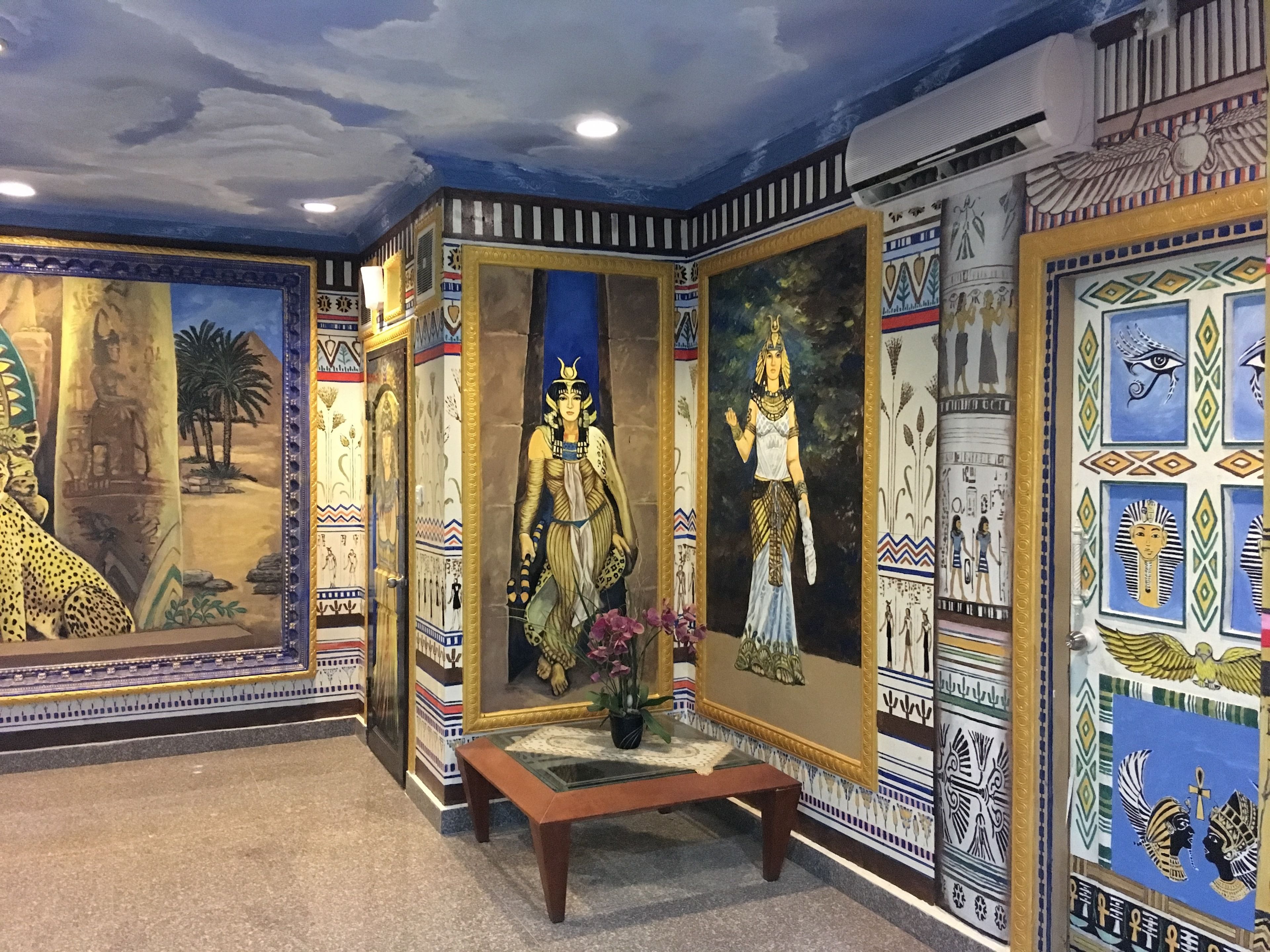 A room with Egyptian-themed murals, a central painting of a figure, and a bench.