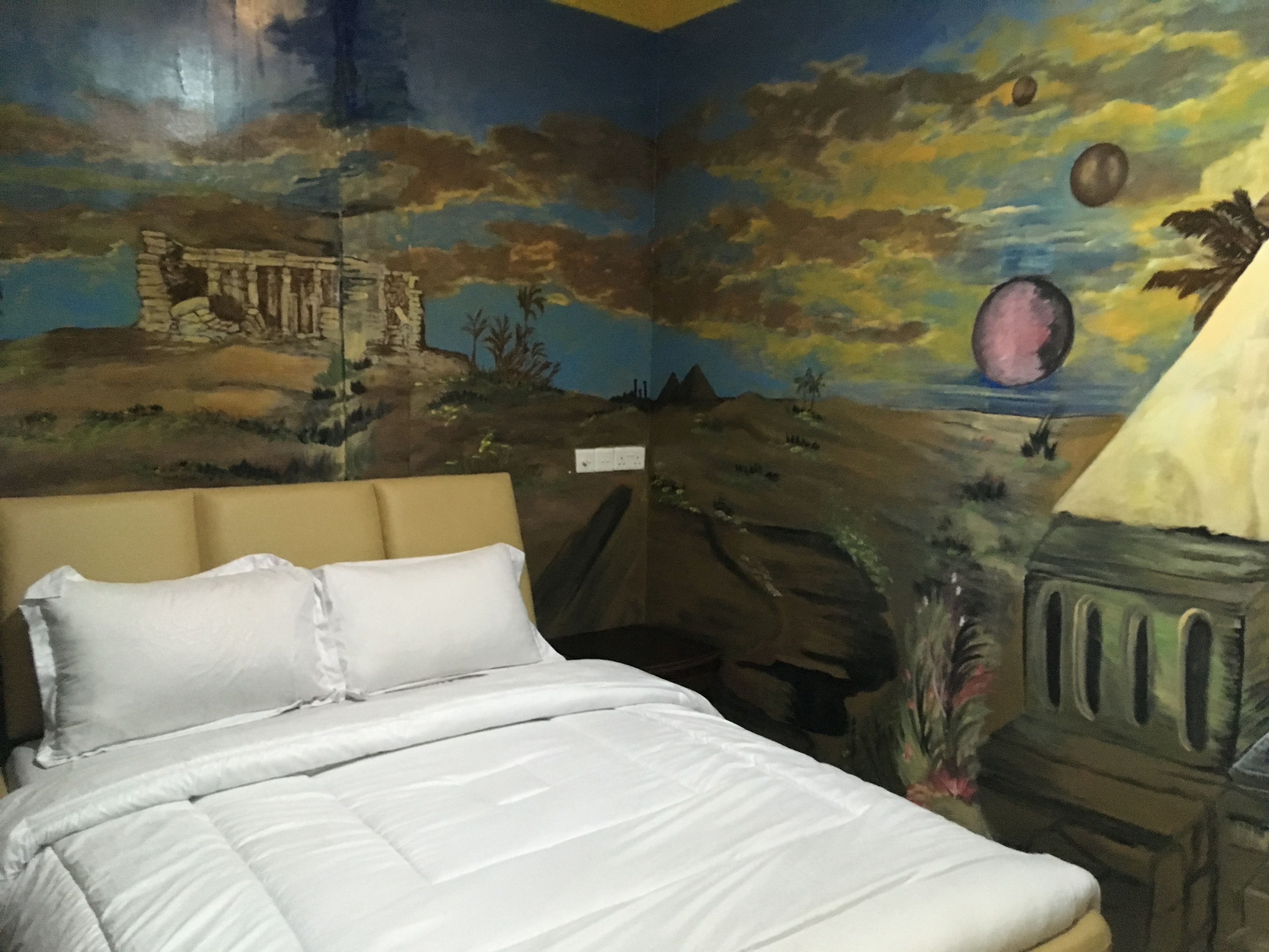 Un dormitorio con cama, cabecera y un mural en la pared que muestra un paisaje con una pirámide y un planeta.