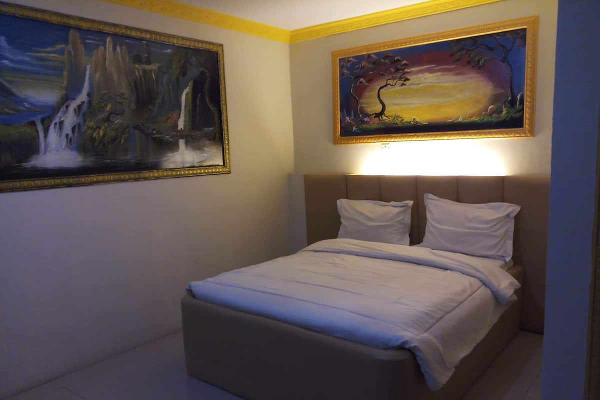 Una cama bien hecha con cabecera, dos almohadas y una colcha. Hay dos cuadros en las paredes, uno con una cascada y el otro con un árbol.
