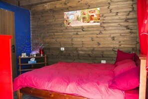 Ein Zimmer mit einer Holzwand, einem Bett mit rosa Bettwäsche, einem kleinen Tisch mit einer roten Lampe und einem gerahmten Bild an der Wand.