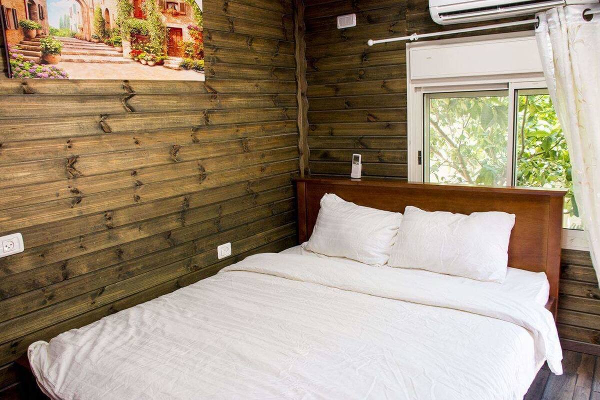 A bedroom with a wooden wall, a bed with white bedding, a window with a view of greenery, and an air conditioning unit.