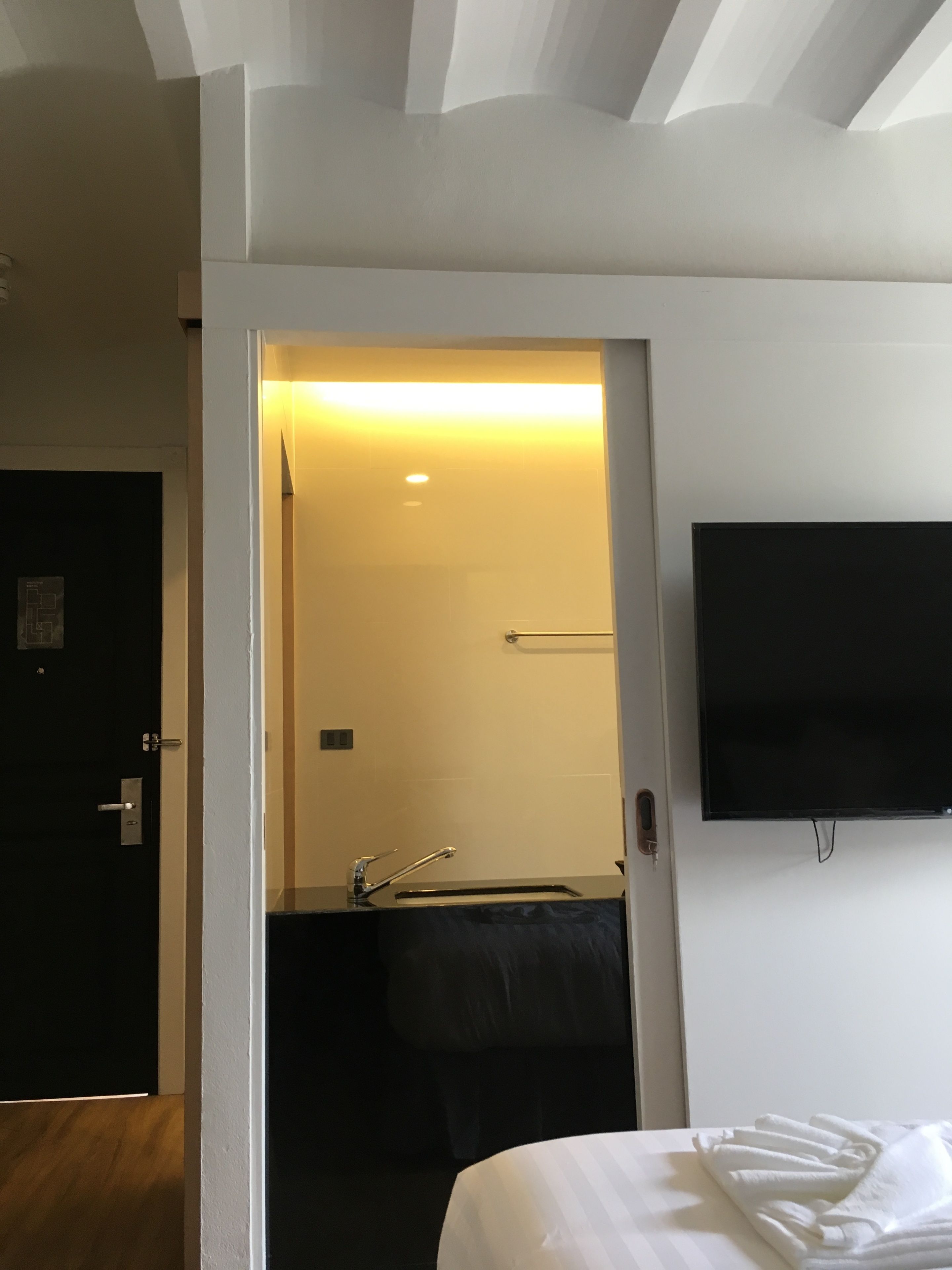 A hotel room with a bed, a TV mounted on the wall, and a bathroom visible through an open door.