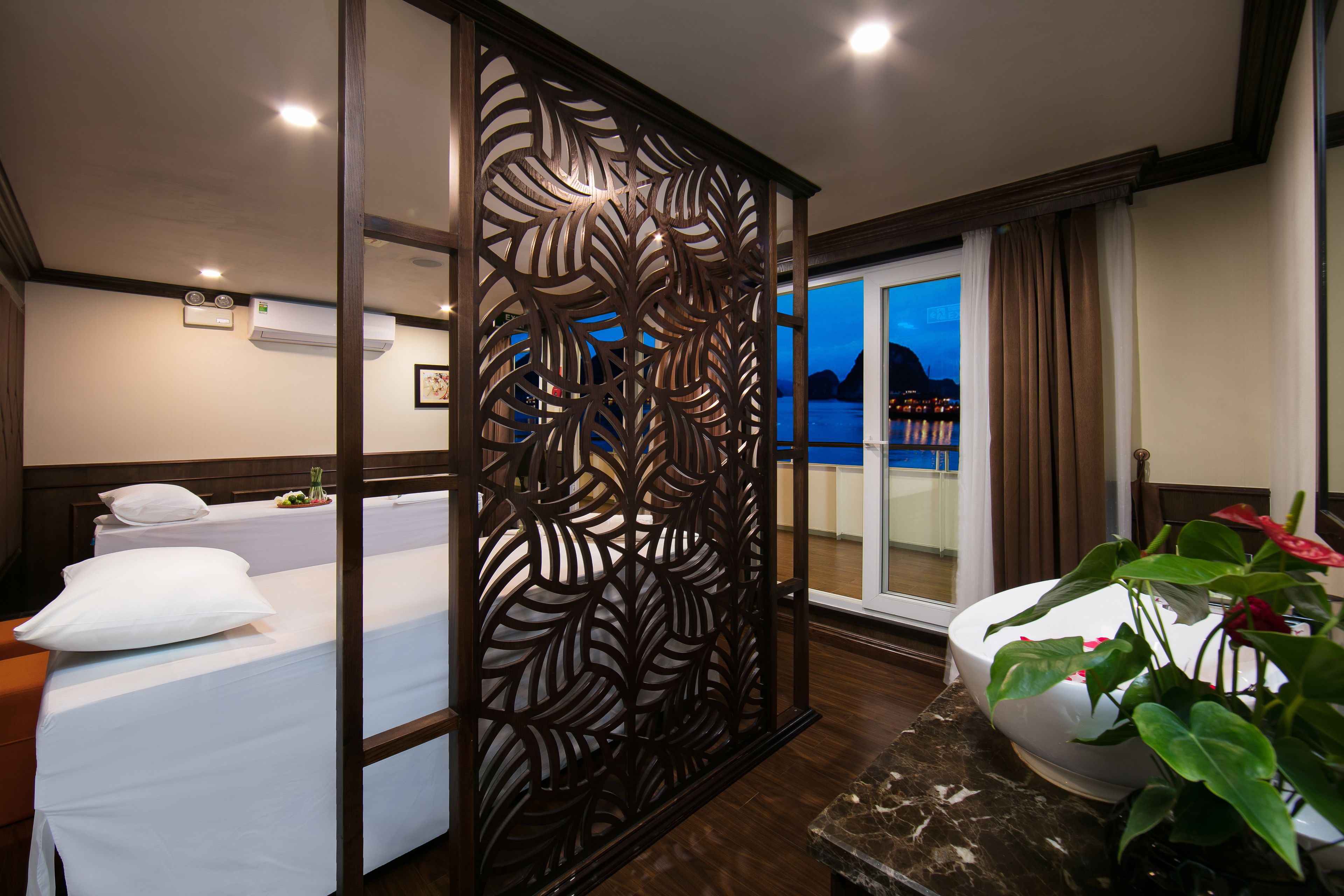 Habitación de hotel con cama, separador decorativo, baño con lavamanos y planta en maceta, y vista a la ciudad al atardecer.