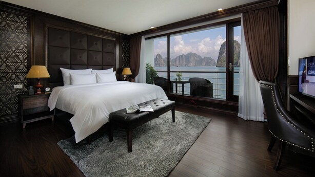 A bedroom with a large bed, two bedside lamps, a bench, a TV, and a view of the sea. - Mon Chéri Cruises (Tuan Chau)