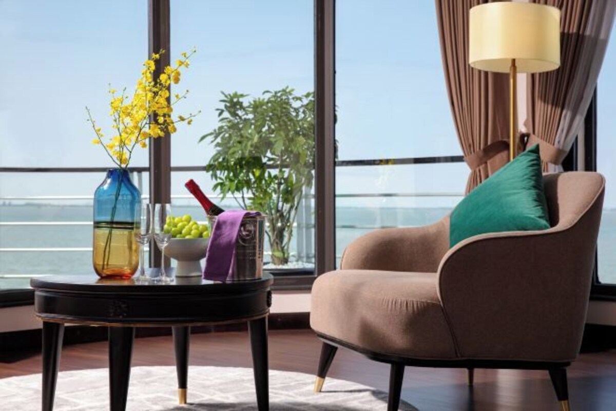 A room with a view of the sea, a chair with a green pillow, a table with a vase of yellow flowers, and a lamp.