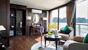 A modern room with a wooden floor, a desk with a mirror, a chair, a round table with a vase of flowers, and a view of the sea through large windows. - Mon Chéri Cruises (Tuan Chau)