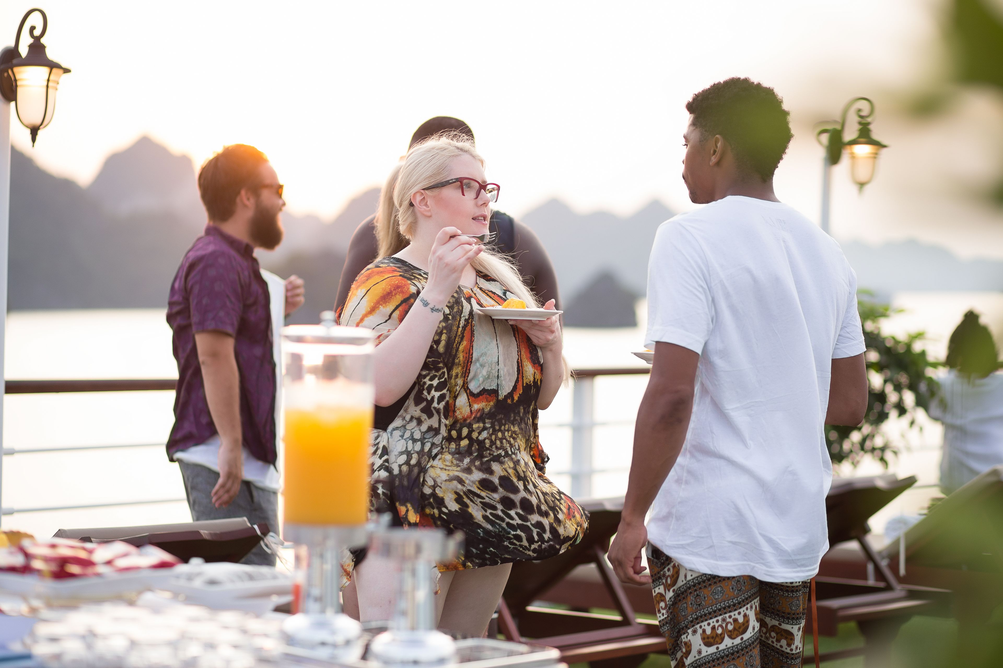 A group of people dining outdoors with a table set for a meal, a drink, and a scenic view in the background.