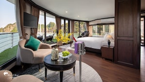A spacious suite with a large bed, a seating area with a view of the sea, and a dining table with a vase of flowers. - Mon Chéri Cruises (Tuan Chau)
