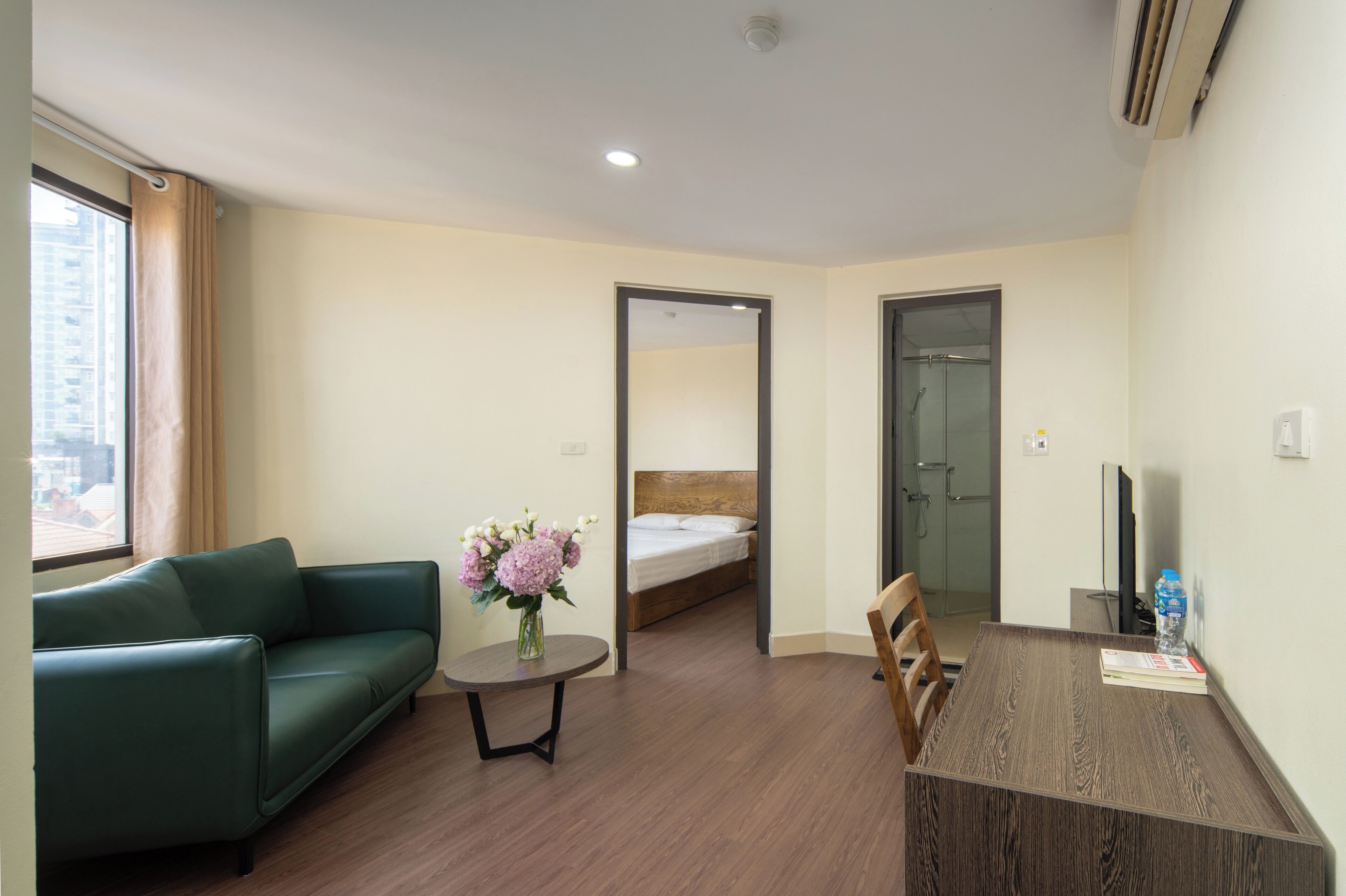 A hotel room with a green sofa, a small table with a vase of flowers, a wooden dining table, a bed, and a TV.