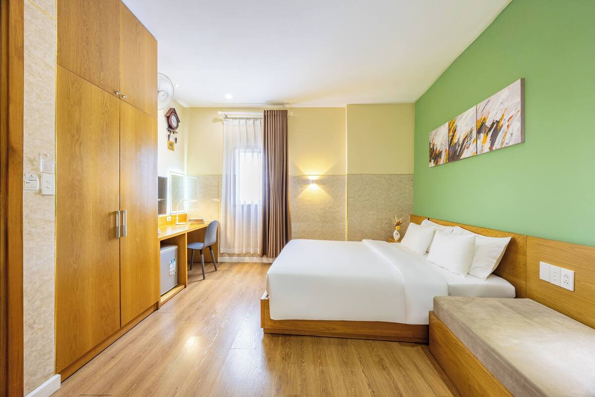 A hotel room with a wooden wardrobe, a desk with a chair, a bed with two pillows, a bedside table, and a wall-mounted artwork.
