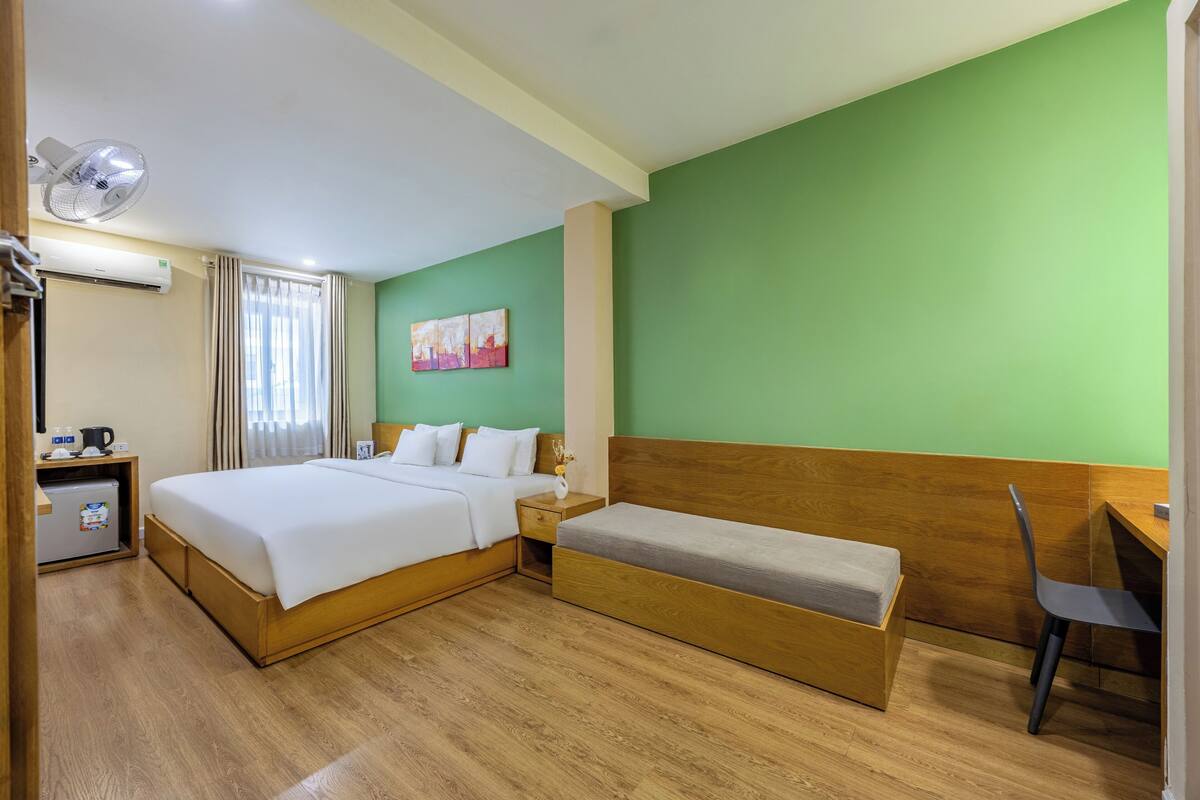 A hotel room with a large bed, a smaller bed, a desk, a chair, a fan, an air conditioner, and a window with curtains.