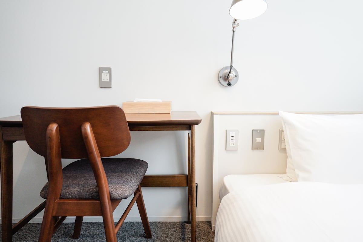 A hotel room with a wooden desk, a chair, a bedside table, a lamp, and a bed.
