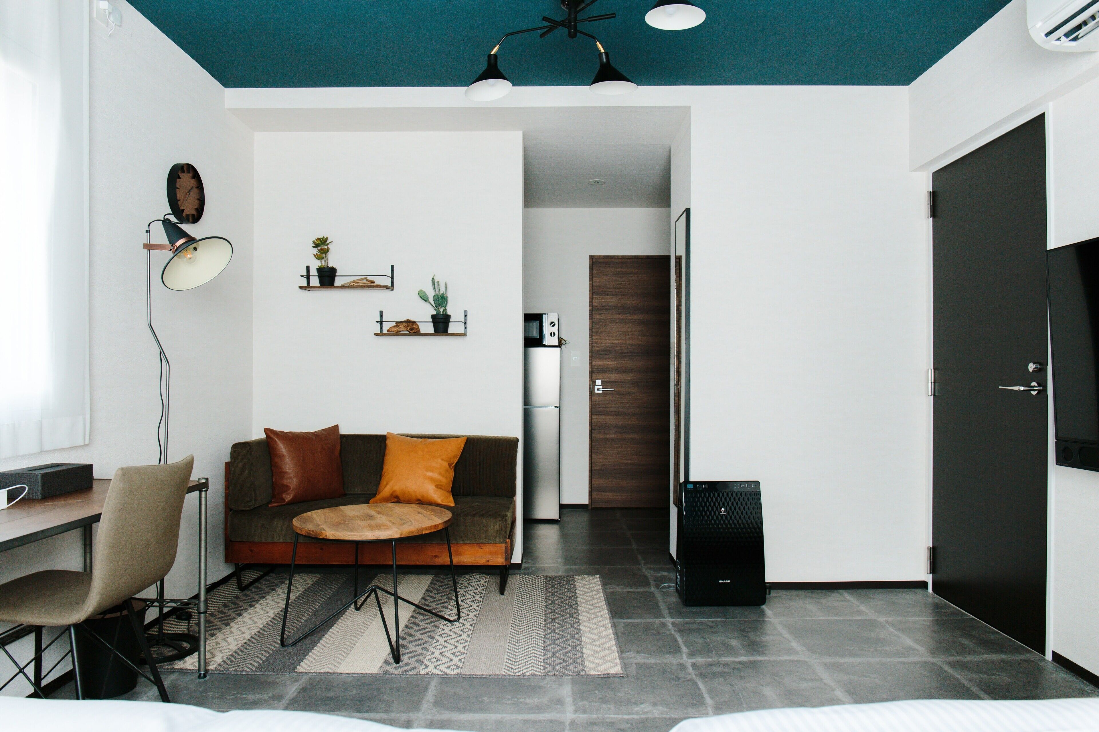A modern living room with a sofa, coffee table, and a desk with a chair. There is a kitchen area with a refrigerator and a microwave.