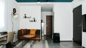 A modern living room with a sofa, coffee table, and a desk with a chair. There is a kitchen area with a refrigerator and a microwave.