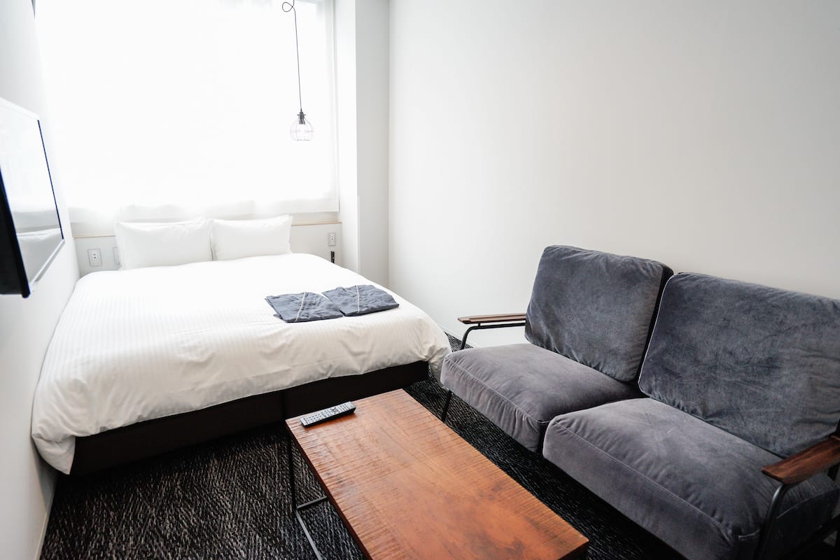 A hotel room with a bed, a grey sofa, a wooden coffee table, and a window with a hanging light fixture.