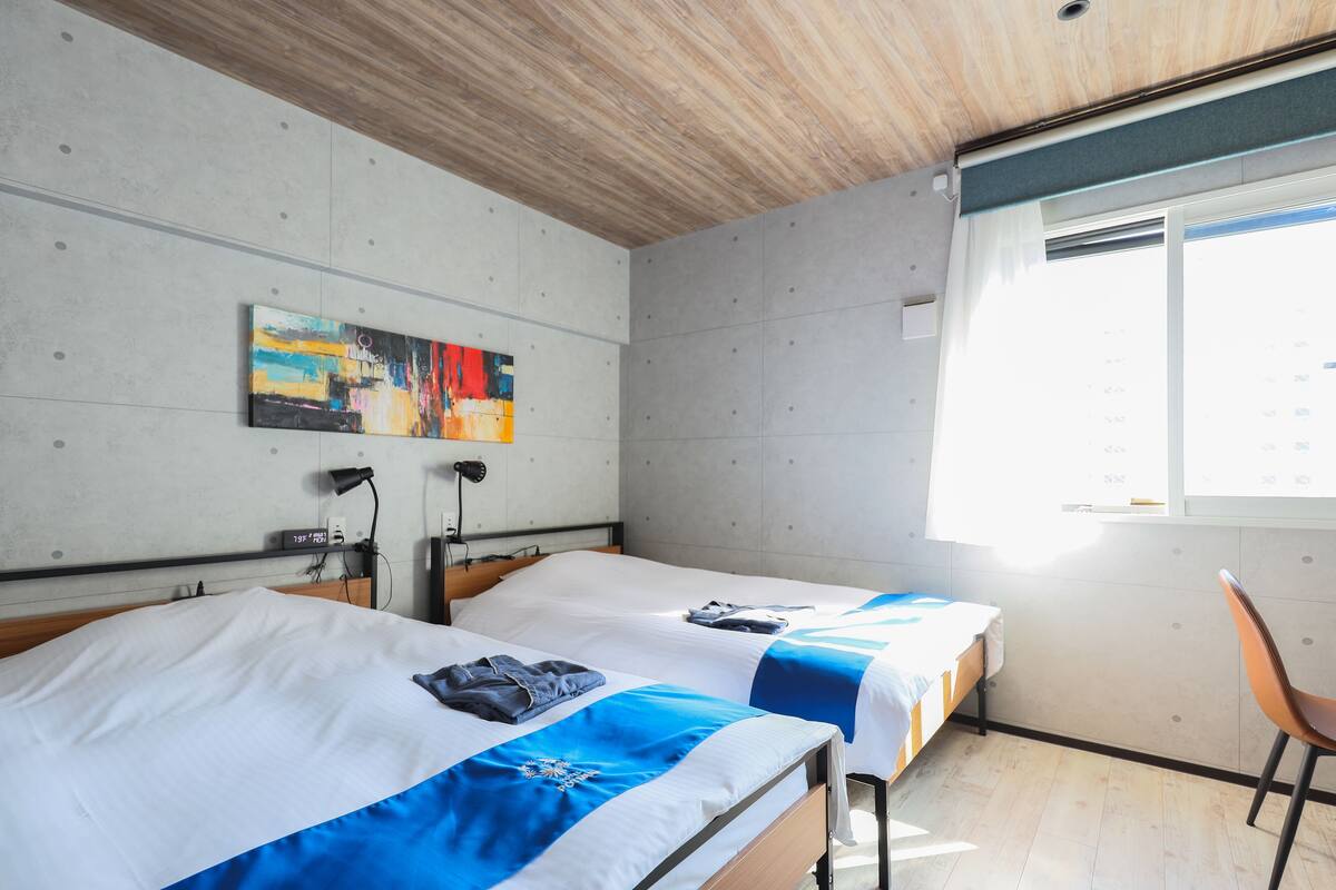 A hotel room with two beds, a painting on the wall, a window with blinds, and a chair.