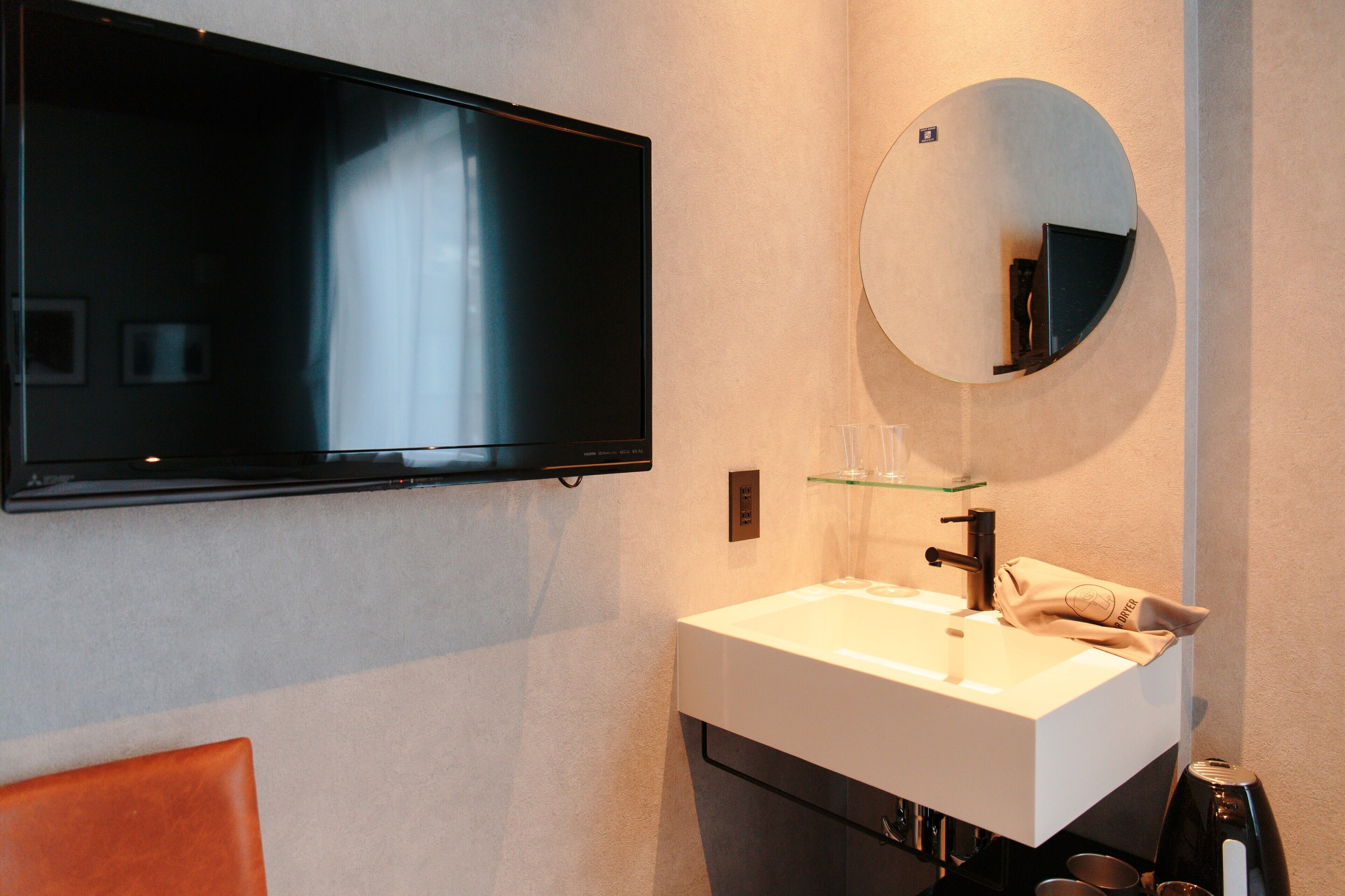 A modern bathroom with a flat-screen TV mounted above a white sink, a round mirror, and a glass shelf.