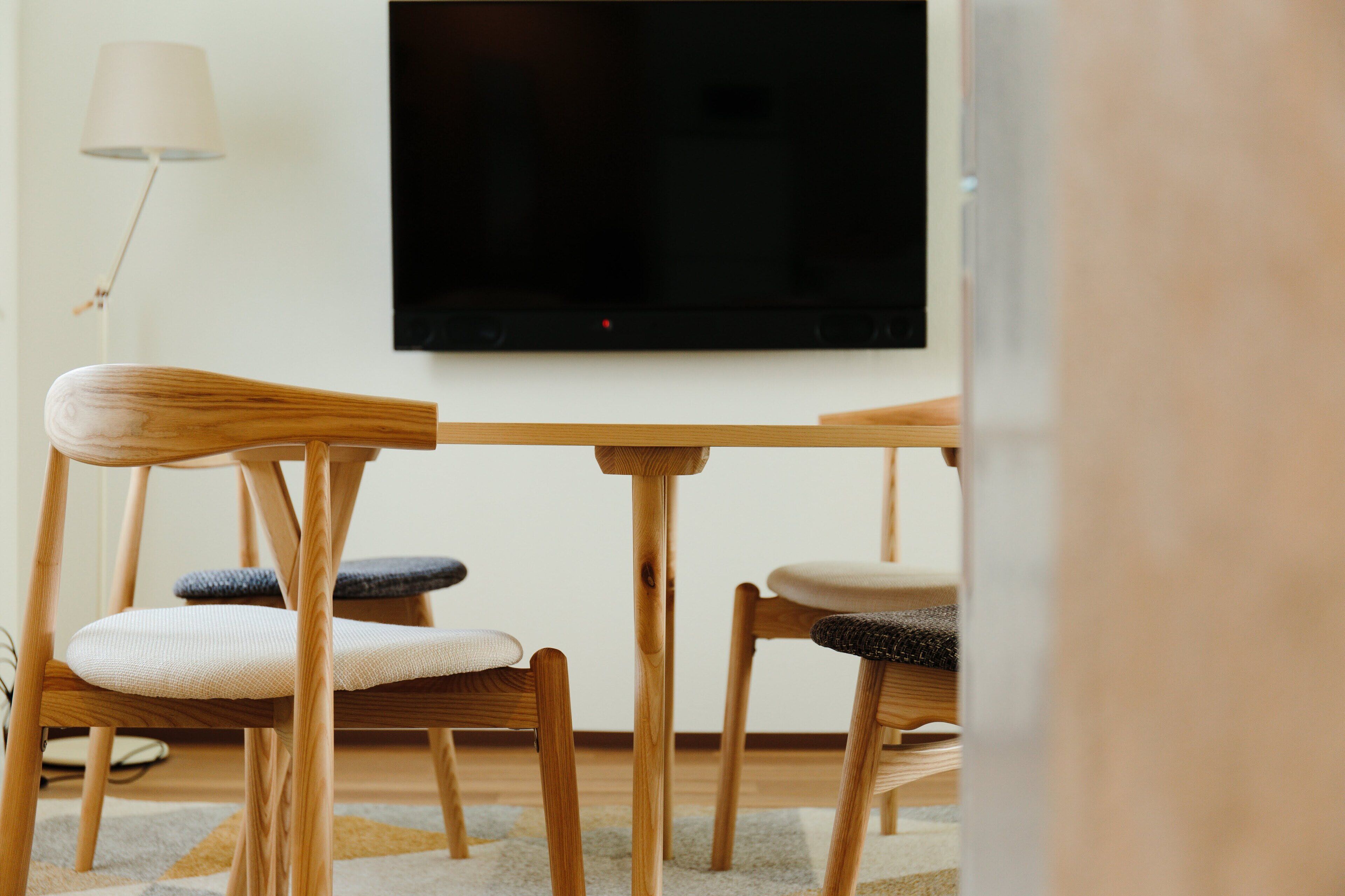 A modern living room with a wooden table and chairs, a flat-screen TV mounted on the wall, and a floor lamp.