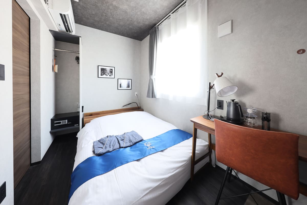 A hotel room with a bed, a desk with a lamp, a chair, and a wardrobe.