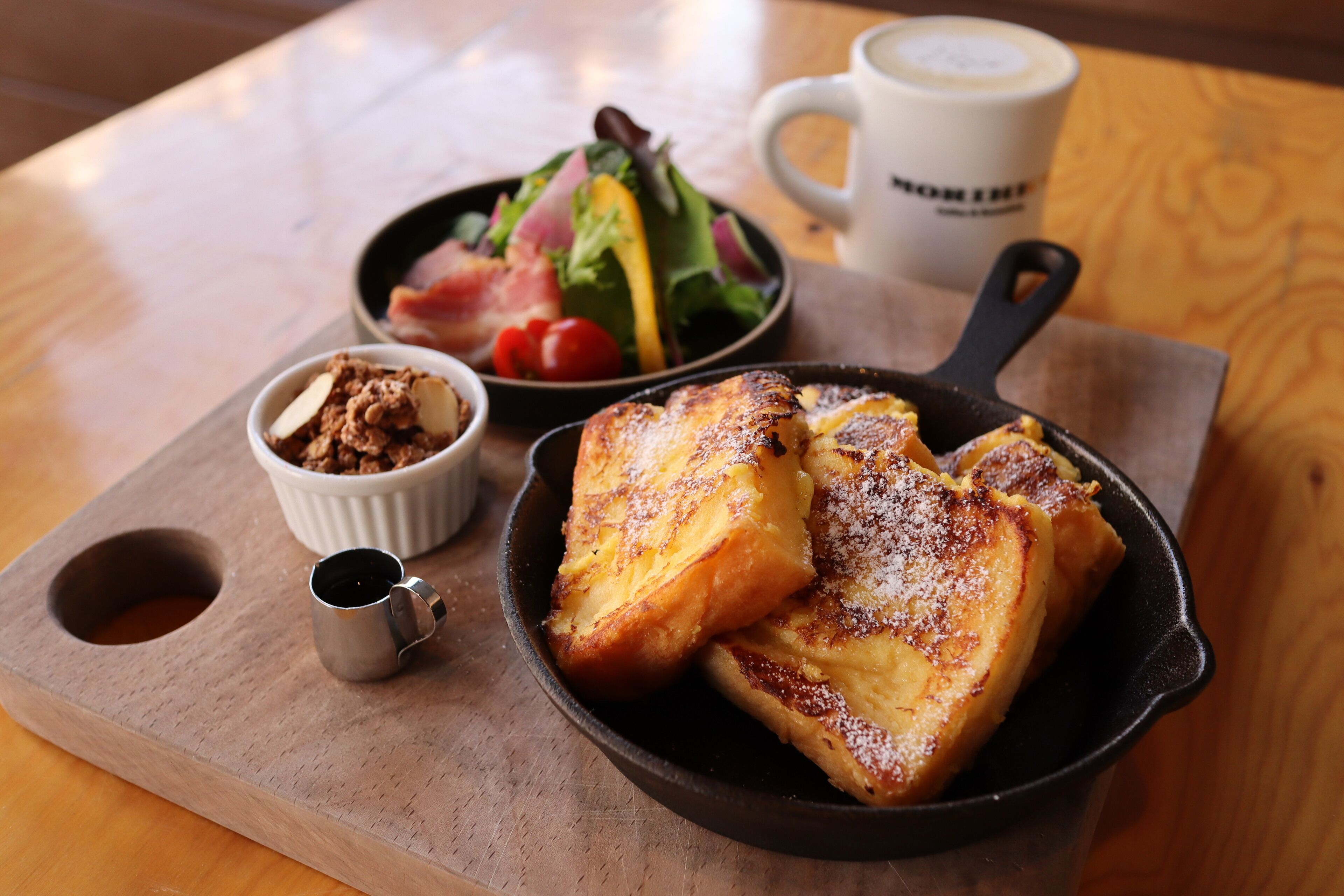 Une table dressée pour le petit-déjeuner avec une poêle contenant des tranches de pain perdu, un saladier rempli de salade, un petit bol de fruits et une tasse de café, le tout posé sur une planche en bois.