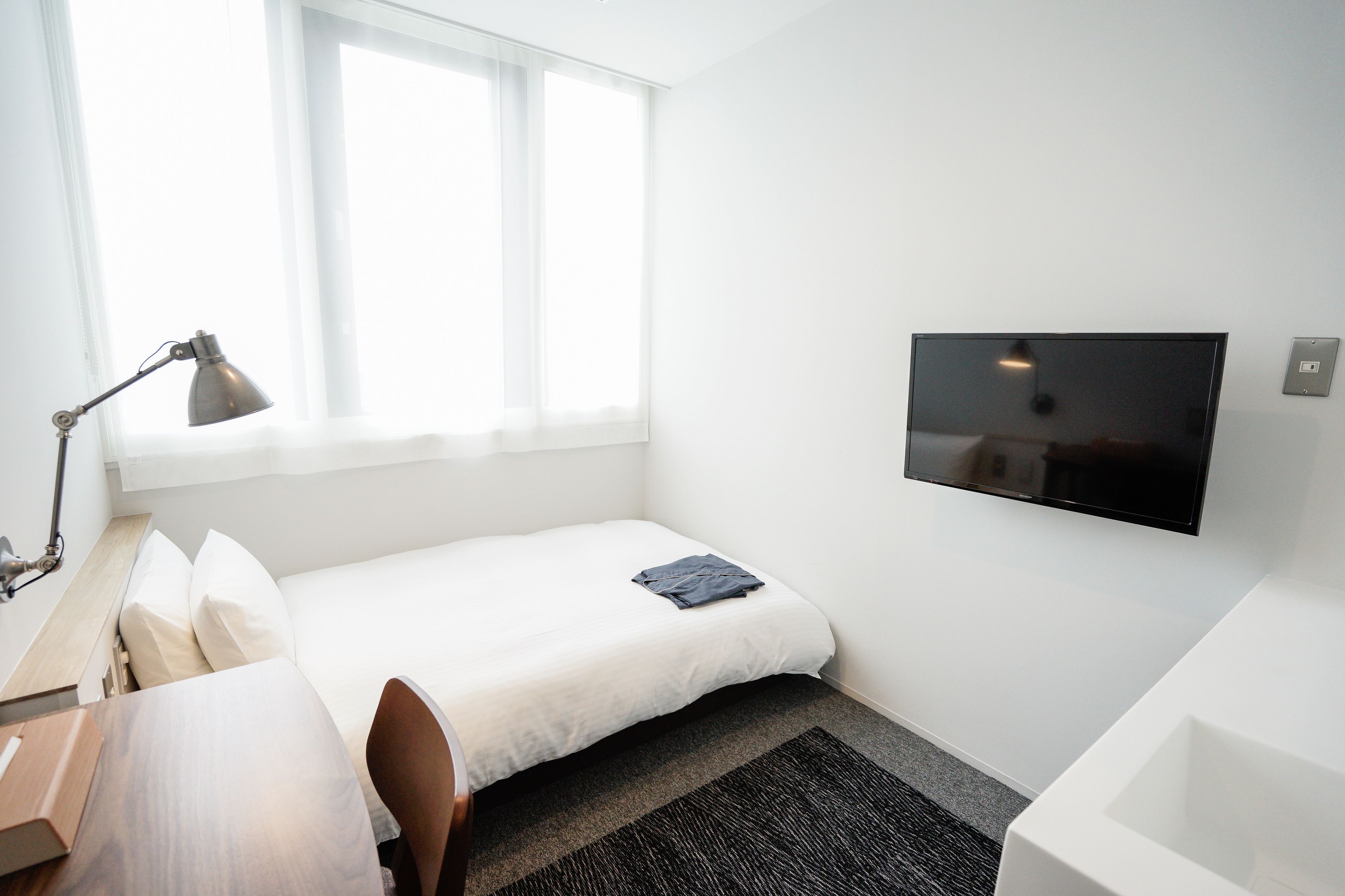 A small, modern hotel room with a single bed, a desk with a lamp, a flat-screen TV mounted on the wall, and a window providing natural light.
