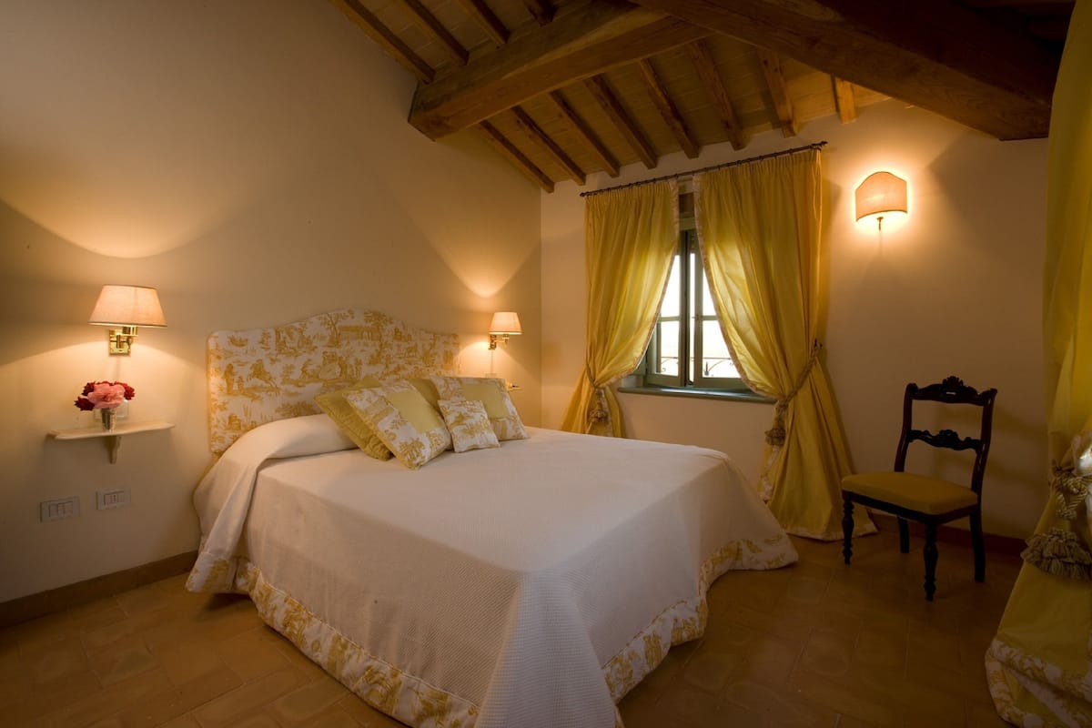Een slaapkamer met een bed, nachtlampjes, een stoel en een raam met gordijnen.