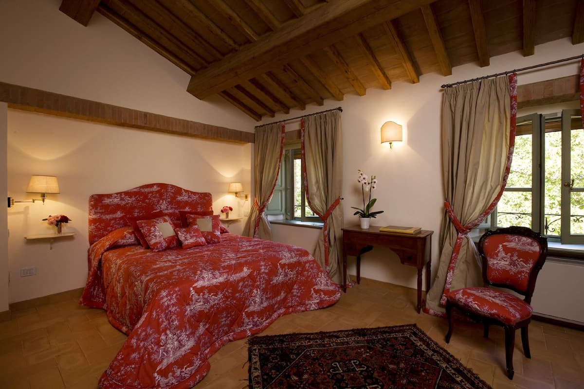Een slaapkamer met een groot bed, een stoel, een tafeltje en een raam met gordijnen.