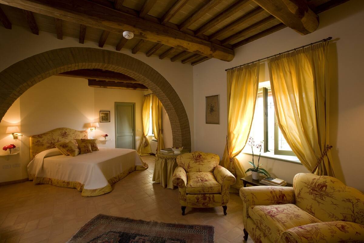 Een slaapkamer met een bed, twee fauteuils, een tafeltje en een raam met gordijnen.