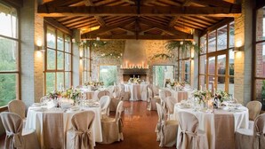 A banquet hall with round tables set for a formal event, featuring a stone fireplace and large windows. - Castello di Gargonza (Monte San Savino)