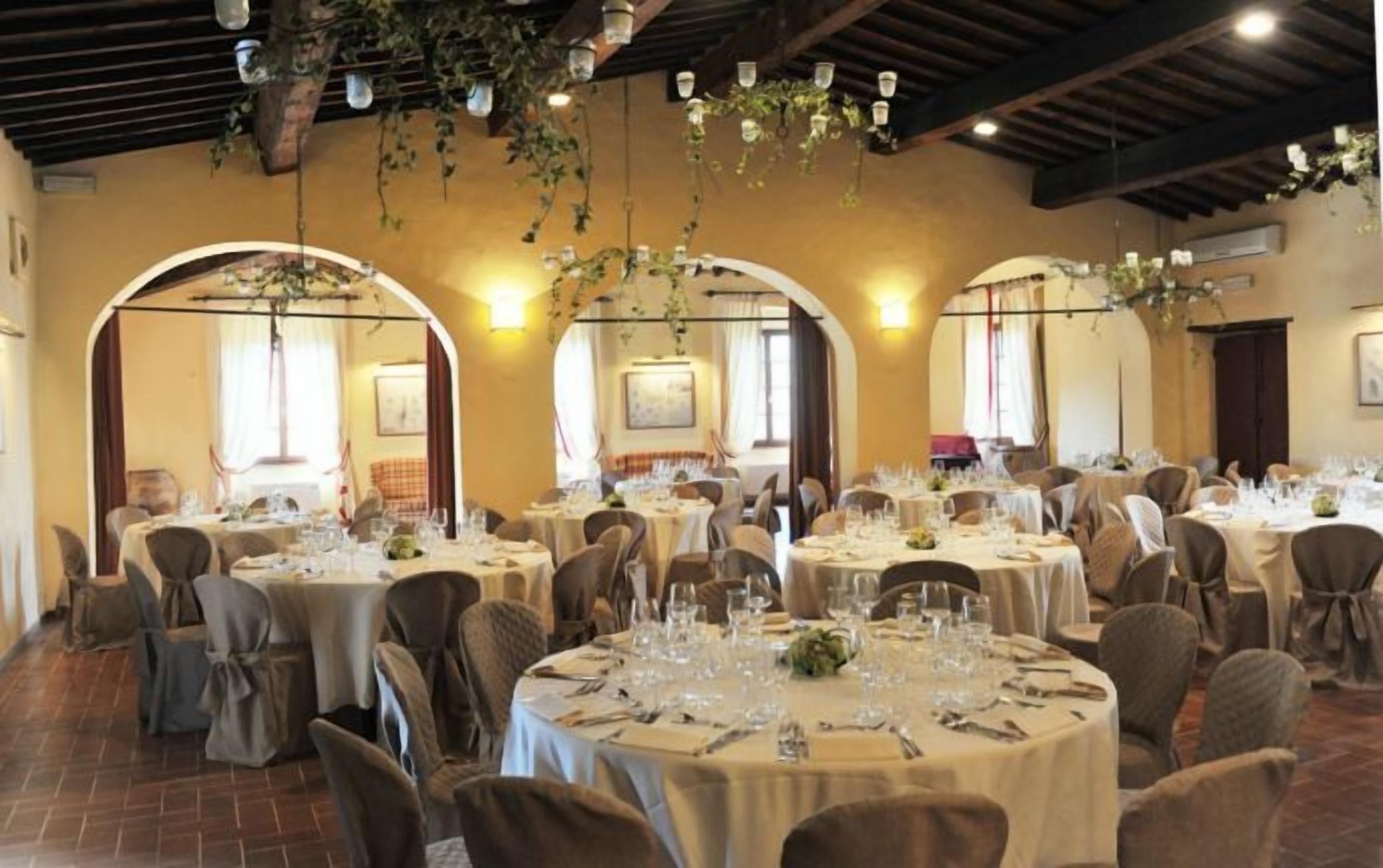 A dining area with round tables set for a meal, featuring a rustic ceiling with hanging plants and exposed beams.