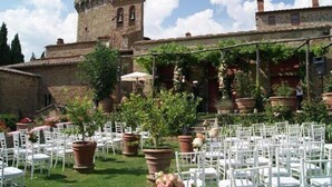 An outdoor seating area with white chairs and tables, surrounded by potted plants and a stone building with a tower. - Castello di Gargonza (Monte San Savino)