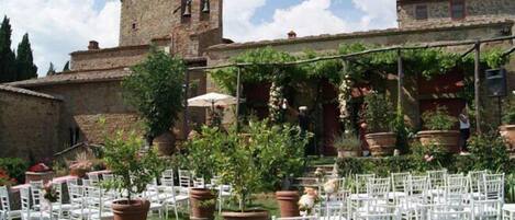 An outdoor seating area with white chairs and tables, surrounded by potted plants and a stone building with a tower.