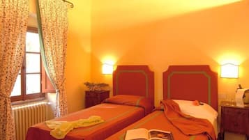Quarto com duas camas, janela com cortinas, luminárias de cabeceira, radiador e uma pequena mesa com uma planta.