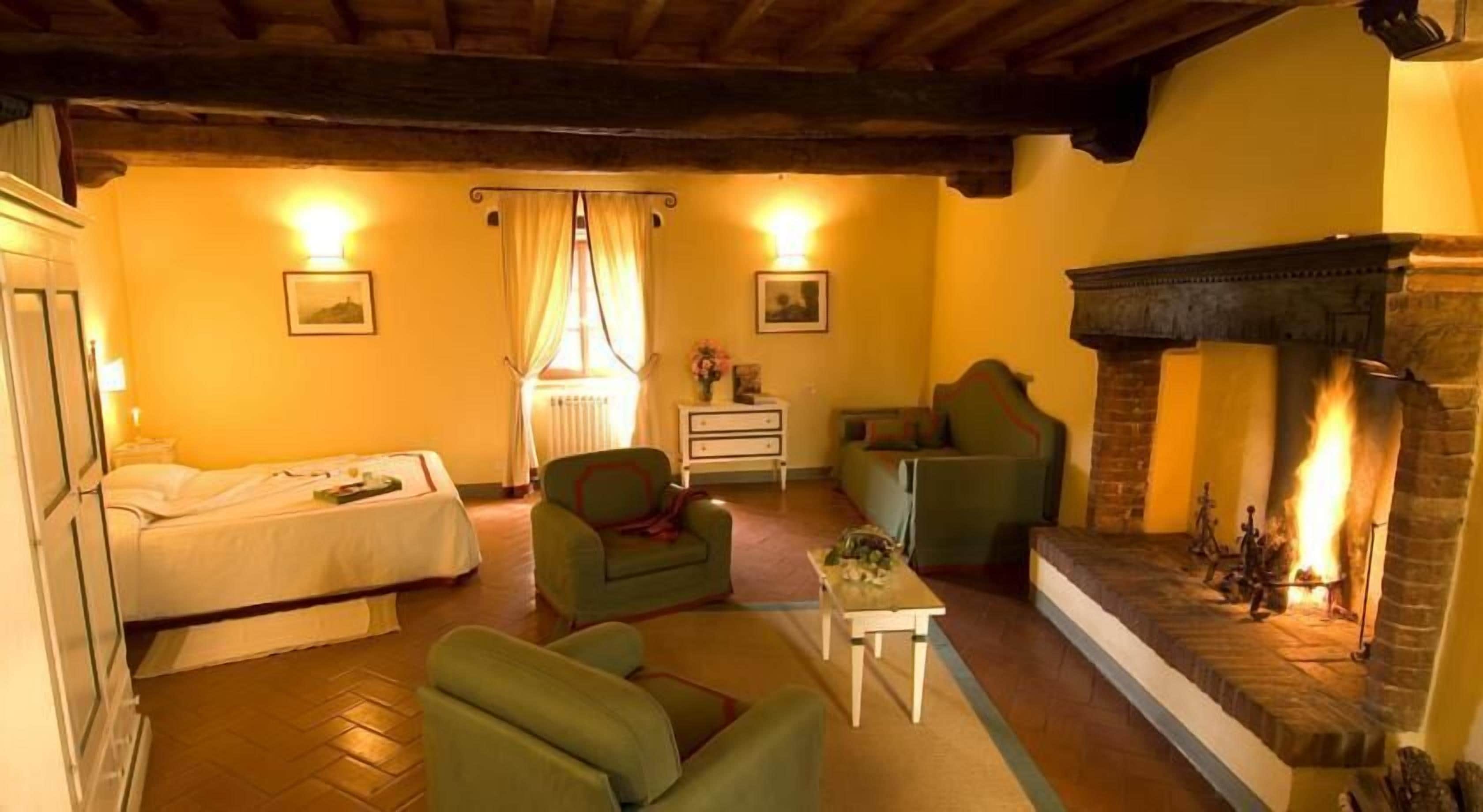 A cozy room with a bed, two green armchairs, a small table, a fireplace, and a nightstand with a lamp.