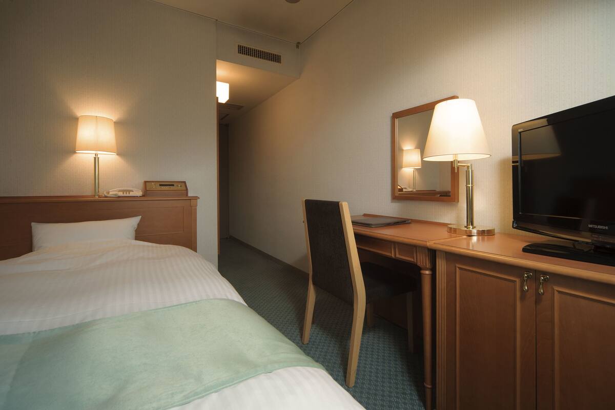 Quarto de hotel com cama, escrivaninha, cadeira, televisão, luminária e telefone na mesa de cabeceira.