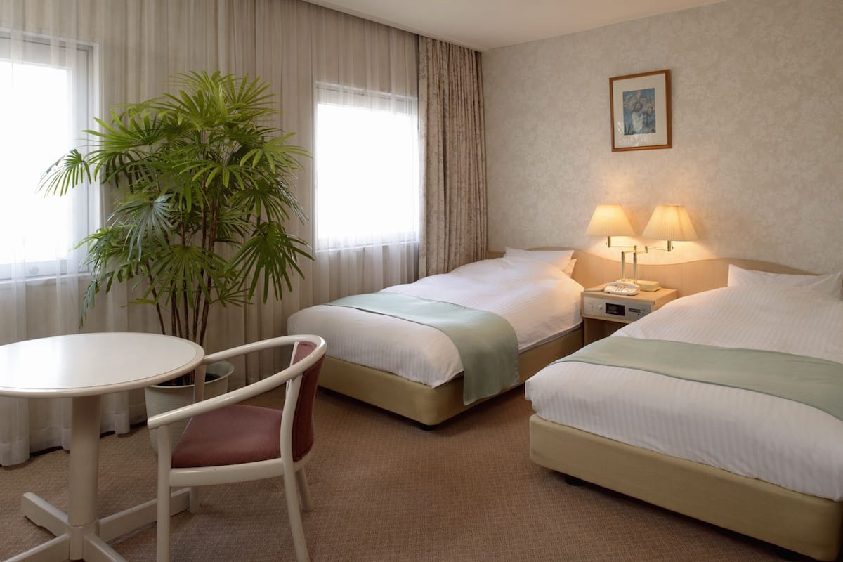 Quarto de hotel com duas camas, uma mesa de jantar, uma cadeira e um vaso de planta.