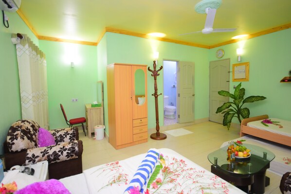 A room with a bed, a sofa, a wooden wardrobe, a small table with a bowl of fruit, a plant, and a ceiling fan. - Olhumathi View Inn (Ukulhas)