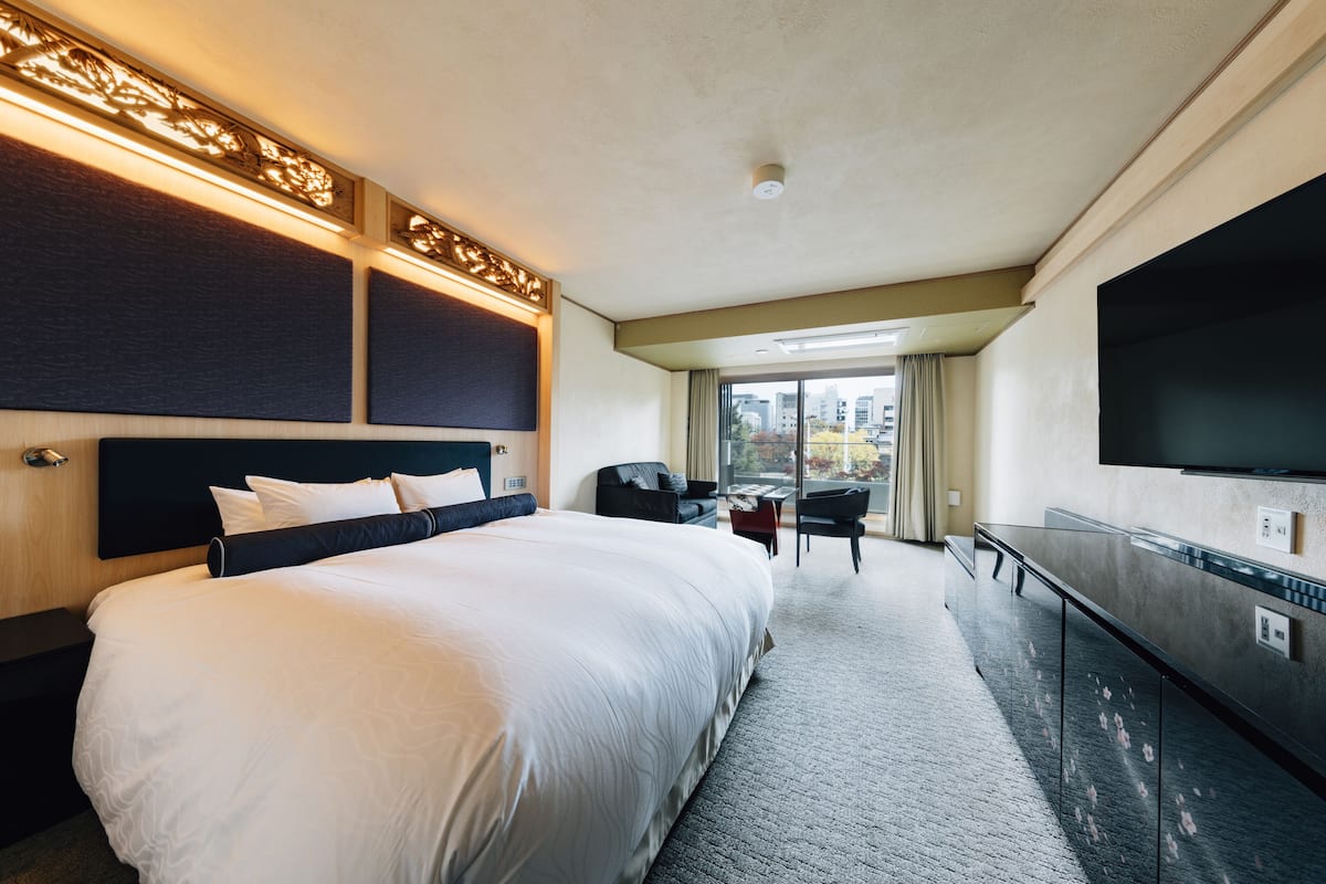 A hotel room with a large bed, a desk, a chair, a flat-screen TV, and a view of the city through the window.